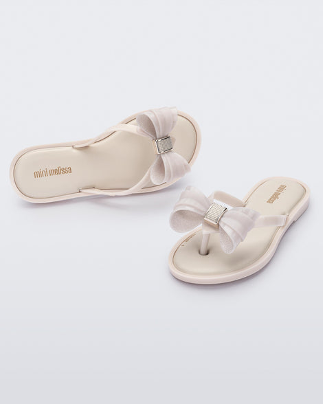 Angled view of a pair of Mini Melissa Flip Flops in beige with bow applique