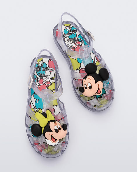 Top view of a pair of clear glitter Mini Melissa Possession + Disney 100 sandal with Mickey on one foot and Minnie on another foot.