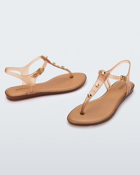 Angled and side view of a pair of clear beige Melissa Solar studs sandals with gold studded t-strap and gold buckle.