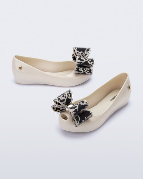 Angled view of a pair of beige kids Ultragirl Classic Bow kids flats with black and clear bow