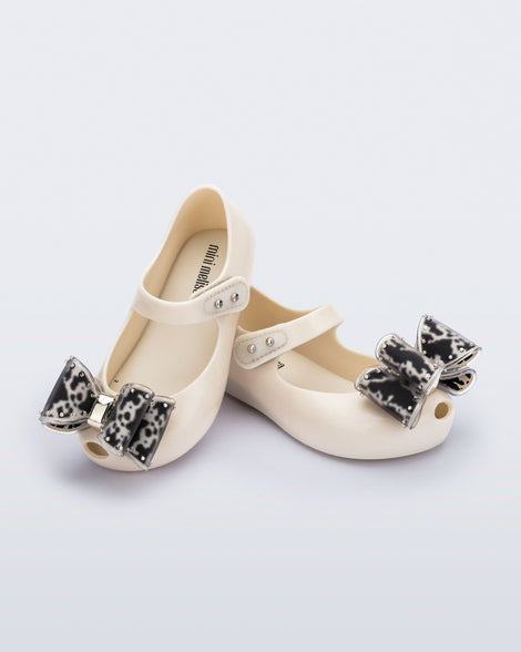 Angled view of a pair of beige baby Ultragirl Classic Bow flats with black and clear bow