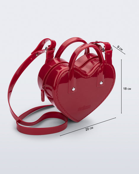 Angled view of a red Heartbeat bag with small handles and longer strap and bag dimensions: 20cm x 9cm x 18 cm.