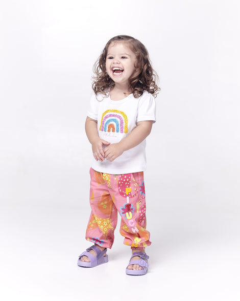 A baby model posing for a picture in a white graphic tee, pink patterned pants and a pair of lilac glitter Mini Melissa Cozy sandals with with two front straps with buckle detail