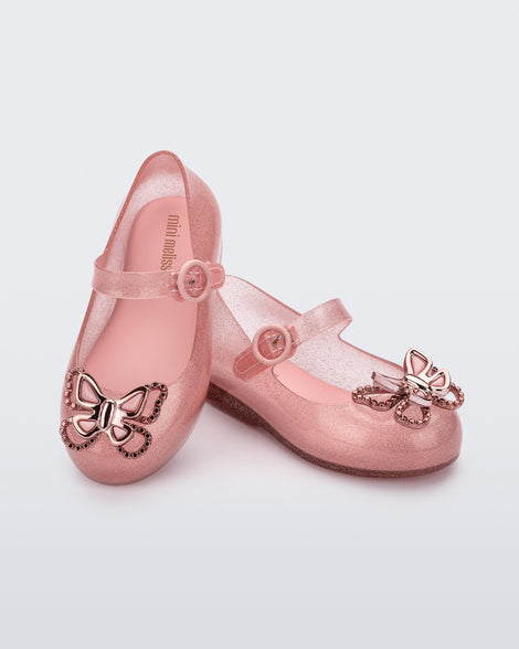 An angled front and top view of a pair of glitter pink Mini Melissa Sweet Love Butterfly flats, leaning on eachother, with a top strap and metallic pink butterfly detail on the toe