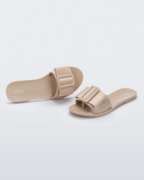 An angled front and top view of a pair of Beige Melissa Babe slides with a buckle like bow detail on the front strap.