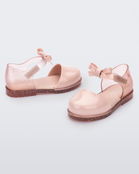 An angled and side view of a pair of pink Mini Melissa Amy sandals with a pink closed toe section and a back pink glitter strap with a lace like bow detail.