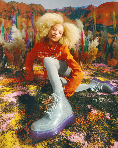 A model sitting on a colorful painted ground with her legs crossed, posing for a picture wearing a pair of lilac/silver glitter Melissa Coturno with a clear silver glitter base, white laces and a lilac sole.