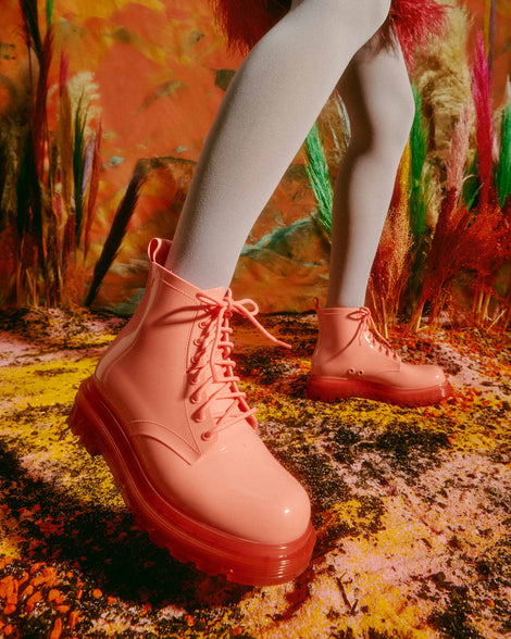 A model's legs wearing a pair of clear pink/pink Melissa Coturno boots with a pink base, laces and sole, standing in front of a painting.