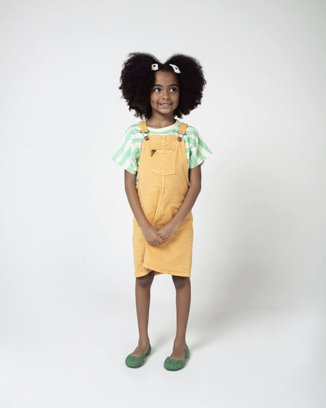 A model posing for a picture in a top and yellow overalls, wearing a pair of green Mini Melissa Campana flats with an open woven texture.