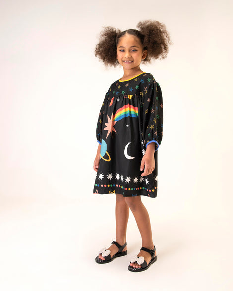 A model posing for a picture in a black patterned dress, wearing a pair of black/beige Mini Melissa Mar Princess sandals with a silver polka dot design insole and a white glitter bow detail on the front strap.