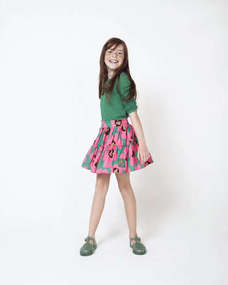 A young model wearing a green top, pink patterned skirt and a pair of Green/Glitter/Silver Mini Melissa Possession sandals with several straps and a green glitter base.