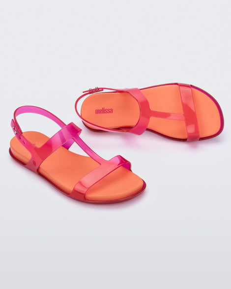 An angled top and side view of a pair of pink / orange Melissa Adore sandals with two translucent pink straps, joined together by a third strap and an orange insole.