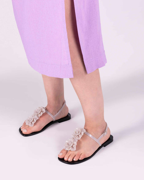 A model's legs wearing a pair of beige/black Melissa Harmonic Squared Garden sandal with a beige flower decoration on the front strap and a black sole.