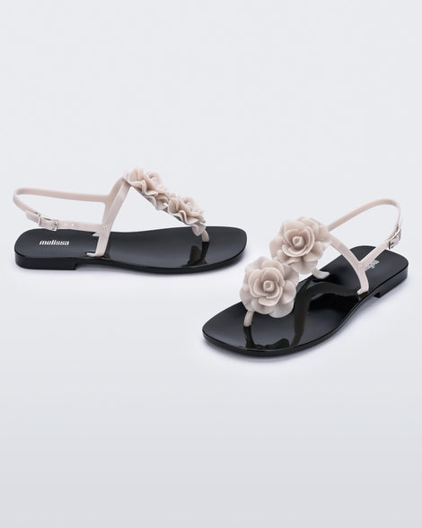 An angled front and side view of a pair of beige/black Melissa Harmonic Squared Garden sandals with a beige flower decoration on the front strap and a black sole.