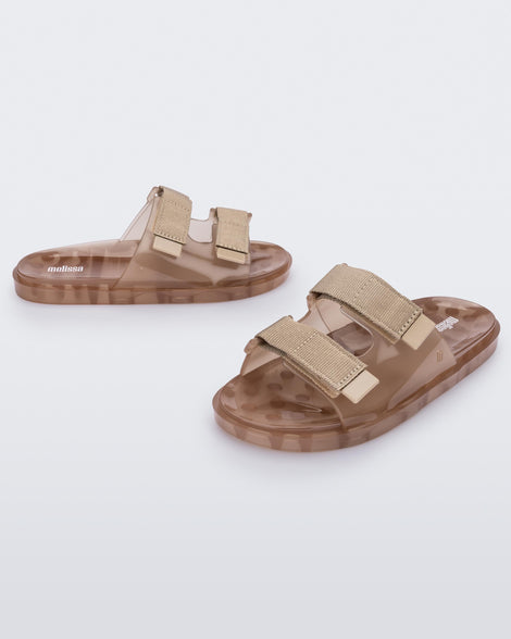 An angled front and side view of a pair of brown Melissa Brave Wide slides with a brown base, velcro straps and a translucent brown insole.