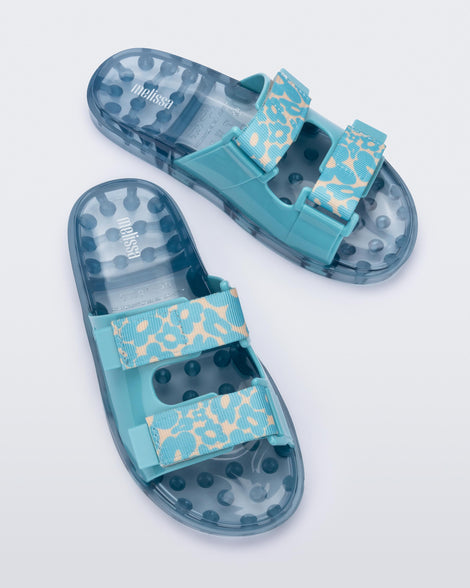 A top view of a pair of blue multicolor Melissa Brave Wide Slides with a blue base, blue and green floral print velcro straps and a translucent blue insole.