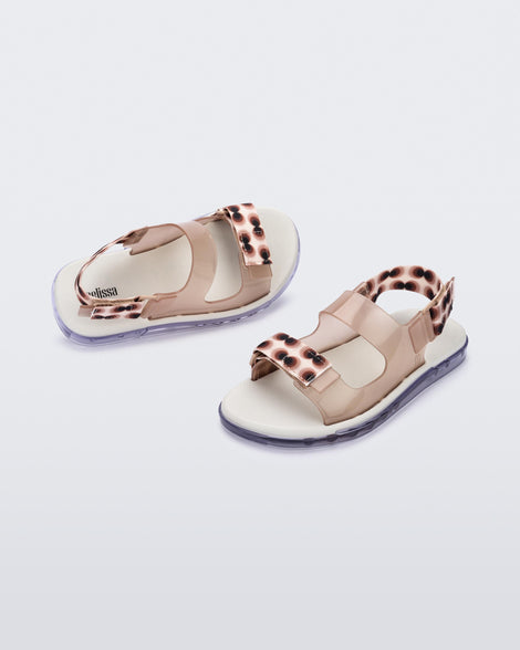 An angled side and top view of a pair of clear brown Melissa Brave Papete sandals with a white insole, brown circle print, transparent beige straps and a velcro back.