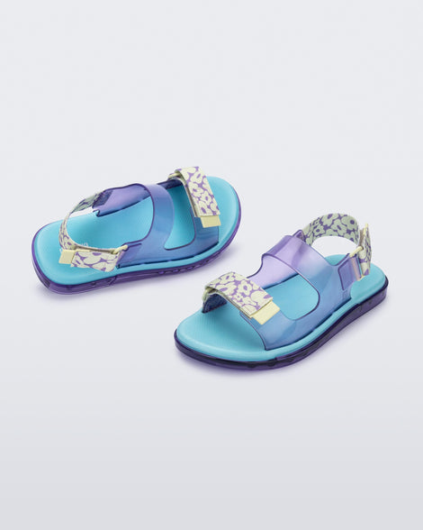 An angled top and side view of a pair of lilac Melissa Brave Papete sandals with a blue insole, purple and green flower print and transparent lilac straps and a velcro back.