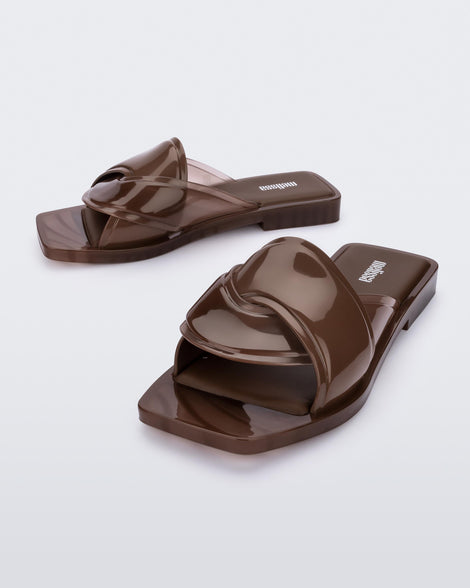 An angled front and side view of a pair of brown Melissa Brigitte slides with a weft designed upper strap.