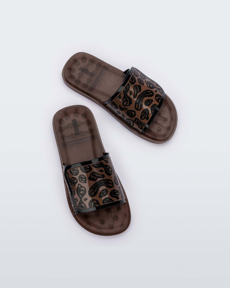 Top view of a pair of brown / black Melissa Brave Slide slides with a black and brown patterned velcro top strap and translucent brown insole.