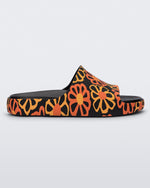 Side view of a multicolor matte mosaic flower Melissa Cloud slide with a black base and an orange and yellow flower pattern, with a black insole reading in orange 