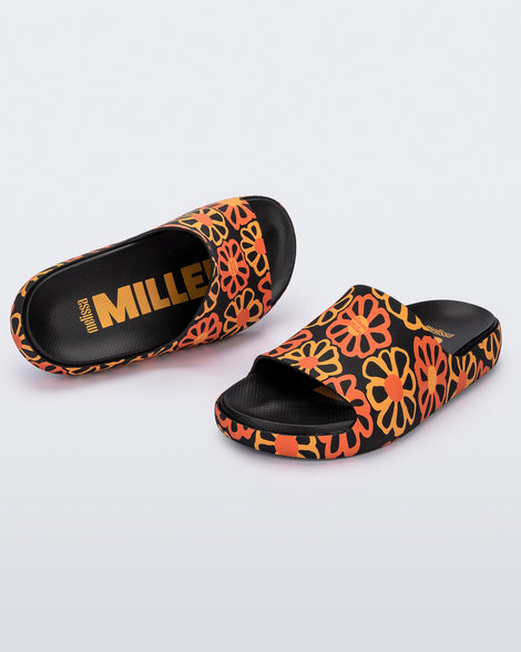 An angled top and front view of a pair of multicolor matte mosaic flower Melissa Cloud slides with a black base and an orange and yellow flower pattern, with a black insole reading in orange 