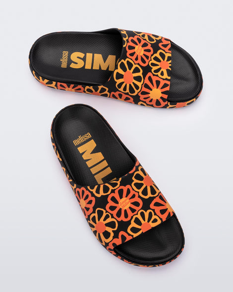 A top and side view of a pair of multicolor matte mosaic flower Melissa Cloud slides with a black base and an orange and yellow flower pattern, with a black insole reading in orange 