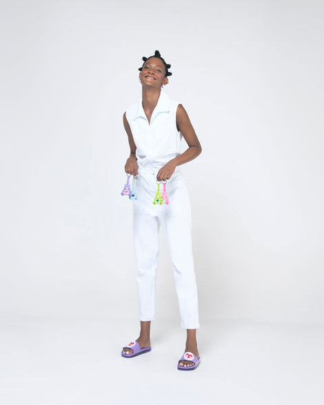 A model posing for a picture in a white top, bottoms, holding 4 care bear keychains and wearing a pair of lilac Melissa Beach slides with 3 pink hearts on a white circle on the top strap, and a care bear patterend lilac insole.