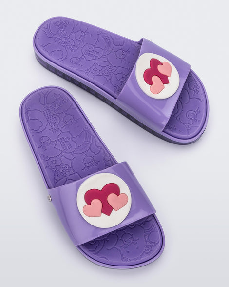 A top view of a pair of lilac Melissa Beach slides with 3 pink hearts on a white circle on the top strap, and a care bear patterend lilac insole.