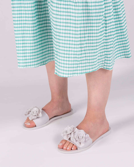 A model's legs wearing green gingham printed dress and a pair of white Melissa Babe Garden slides with two flowers on the front strap.