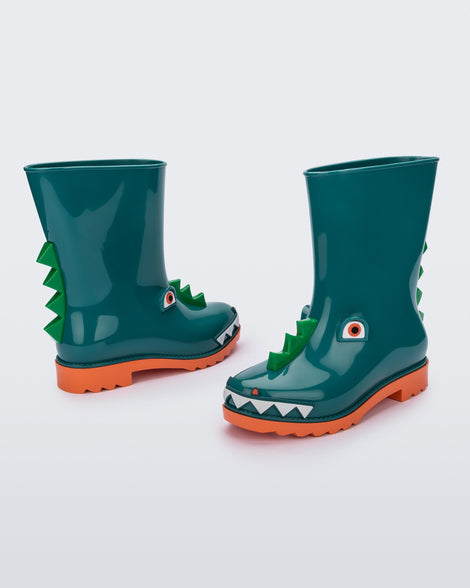 An angled front, back and side view of a pair of orange/green Mini Melissa Rain Boots with a green base, orange sole, light green triangle details, white triangle details in the front and a set of orange eyes which looks like a dinosaurs face,
