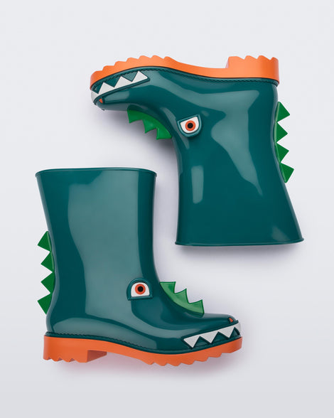 An angled side view of a pair of orange/green Mini Melissa Rain Boots with a green base, orange sole, light green triangle details, white triangle details in the front and a set of orange eyes which looks like a dinosaurs face, laying on their side.