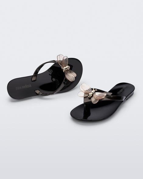 An angled front and top view of a pair of black / metallic black Mini Melissa's Harmonic Bugs flip flop with a clear glitter bug with a gold buckle on the straps.