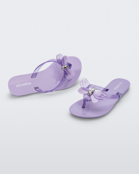 An angled front and top view of a pair of lilac / lilac glitter Mini Melissa Harmonic Bugs flip flops with a clear glitter bug with a silver buckle on the straps.