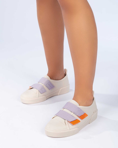 A model's legs wearing a pair of matte beige/lilac Melissa Cool sneakers with two orange and lilac velcro straps.