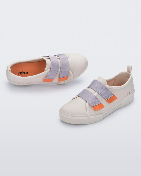 An angled side and top view of a pair of matte beige/lilac Melissa Cool sneakers with two orange and lilac velcro straps.