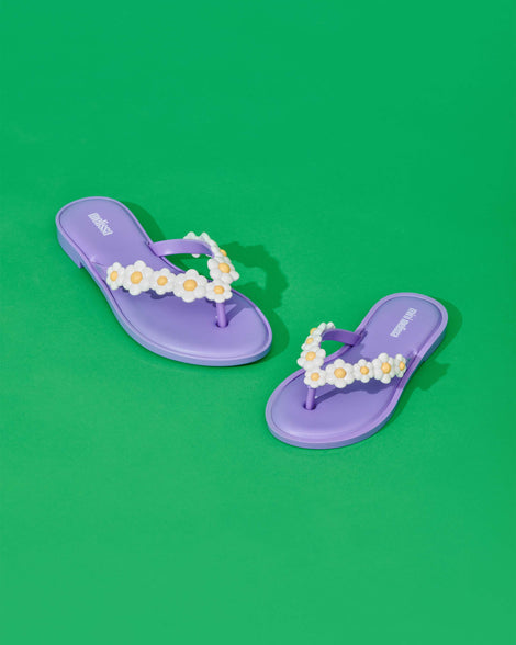 A top and angled view of a pair of lilac / white Melissa Spring Flip Flops with white and yellow flowers on them on a green surface.