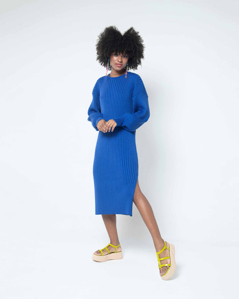 A model posing for a picture in a blue dress and a pair of beige/green Melissa Caribe High Platform sandals with two green front straps joined by a vertical strap intersecting the ankle strap and a beige sole.