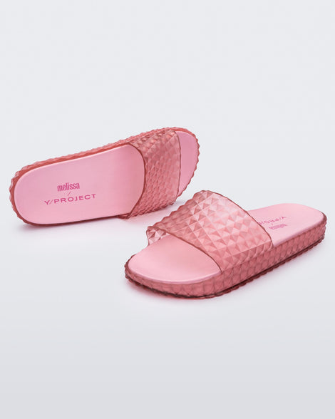 An angled front and side view of a pair of transparent pink Melissa Court Slides with a checkered pattern texture.