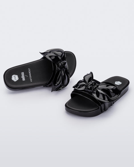 An angled front and top view of a pair of black Melissa Tie Beach slides with a black base and a bow on the front strap.