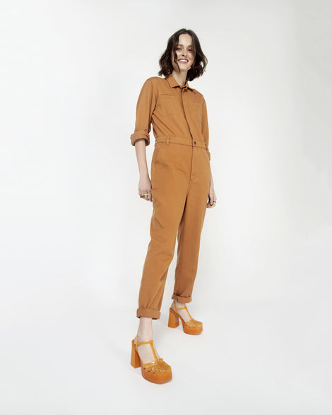 A model posing for a picture in a brown jumpsuit and a pair of orange Melissa Party Heels with several straps and a closed toe front.