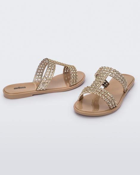 An angled front and side view of a pair of beige / gold Melissa Glowing slides with gold gem stones on the straps and a beige sole.