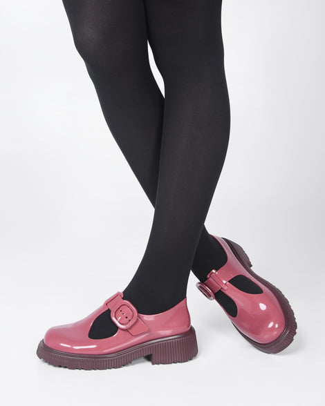A model's legs in black tights, wearing a pair of Burgundy Mini Melissa Jackie loafers with a Burgundy base, two cut outs, a Burgundy buckle detail strap and brown sole.
