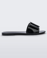 An outter side view of a black Melissa Babe slide with a buckle like bow detail on the front strap.