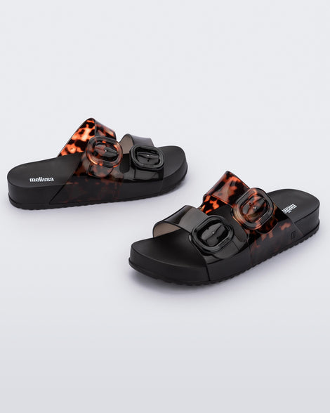 Side view of a pair of Black/Clear Tortoise Melissa Cozy slides with two black and transparent tortoise straps with a buckle detail.