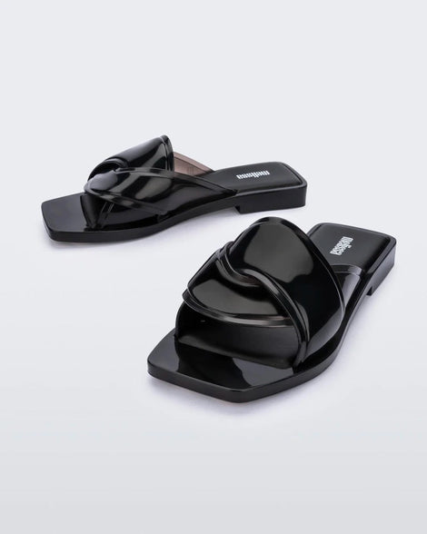 An angled front view of a pair of black Melissa Brigitte slides with a weft designed upper strap.