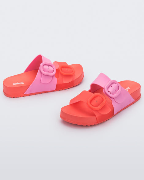 An angled front and side view of a pair of Red/Pink Melissa Cozy slides with two red and pink straps with a buckle detail.