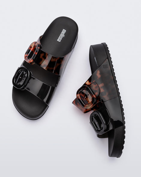 Top and side view of a pair of Black/Clear Tortoise Melissa Cozy slides with two black and transparent tortoise straps with a buckle detail.