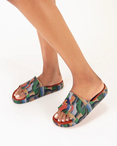 A model's legs wearing a pair of tan multicolor Melissa Cloud slides with an abstract painting design of a house, person, and farmland.