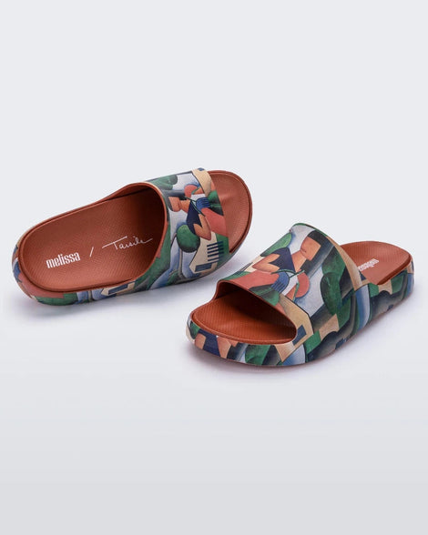 An angled front and top view of a pair of tan multicolor Melissa Cloud slides with an abstract painting design of a house, person, and farmland.